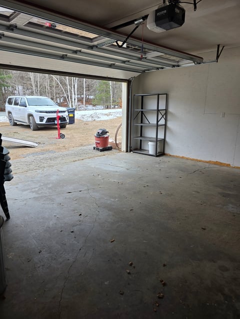 Empty garage interior with open door revealing parked white SUV and wooded driveway outside
