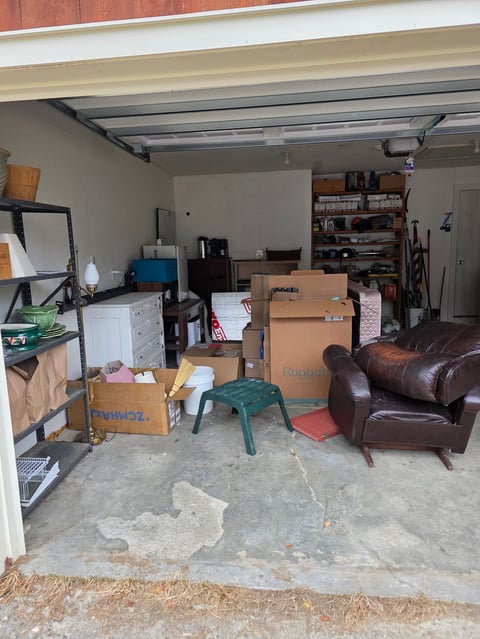 Cluttered garage interior with cardboard boxes, brown leather furniture, tools, and various stored items scattered across the concrete floor