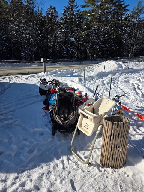 Snowmobile parked on frozen lake with winter gear and trash bin, evergreen forest and snow-covered landscape in background