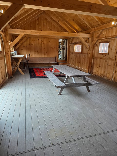 Interior of a wooden barn with exposed beams, wooden floors, a picnic table, small desk area, and red geometric rug