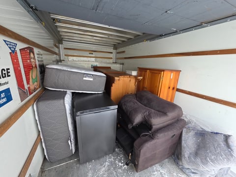 Storage unit interior with stacked mattresses, brown furniture, refrigerator, and boxes along walls with wooden beams