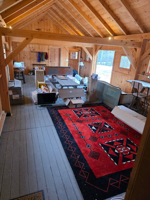 Interior of a wooden cabin with exposed beam ceiling, red patterned rug, bed, desk with computers, and various furnishings