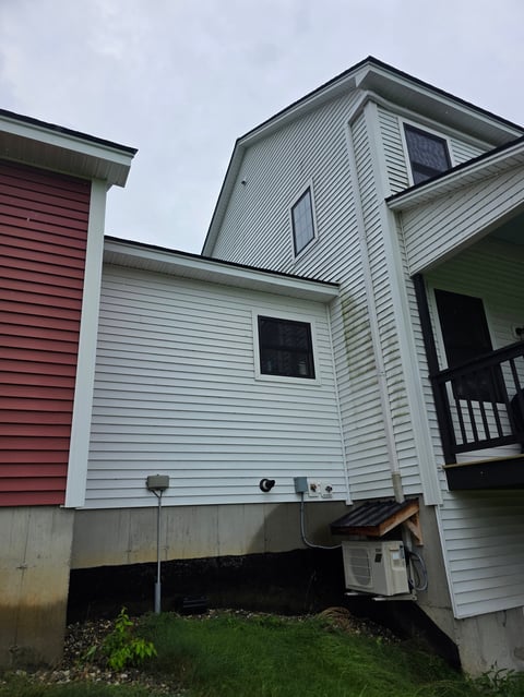 Two-story house with white and red vinyl siding, second-story bay window, front porch deck, and concrete foundation.